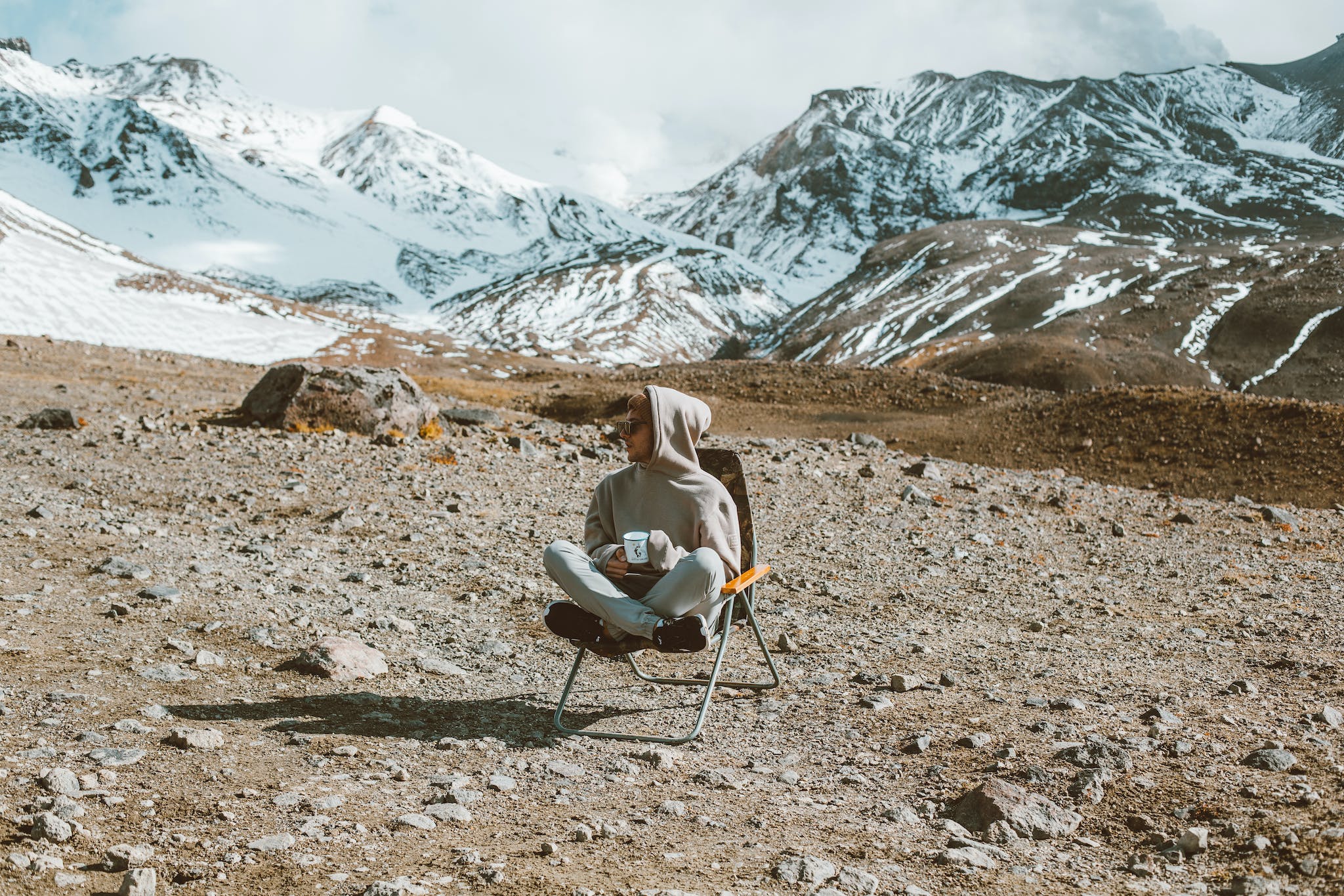 Full body of man sitting on folding chair and drinking tea with high mountains covered with snow on background